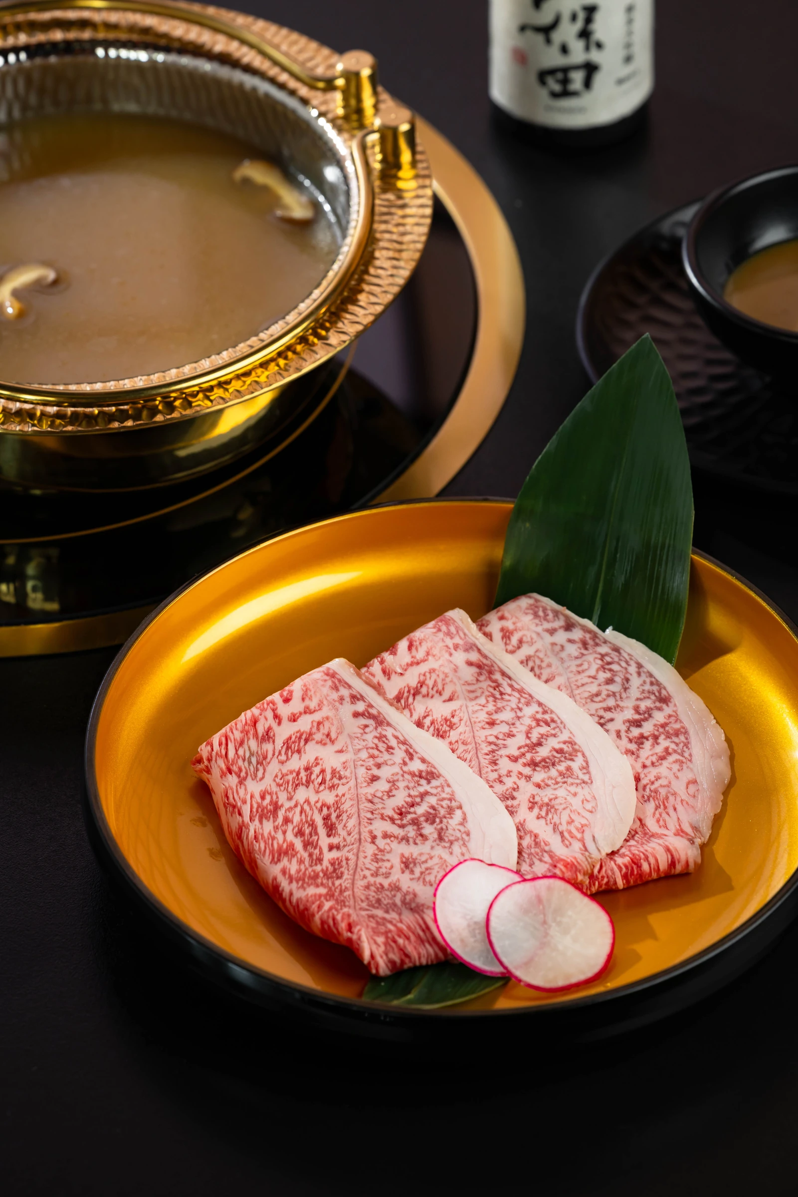 Three round marbled Wagyu slices on a gold-toned plate beside hotpot