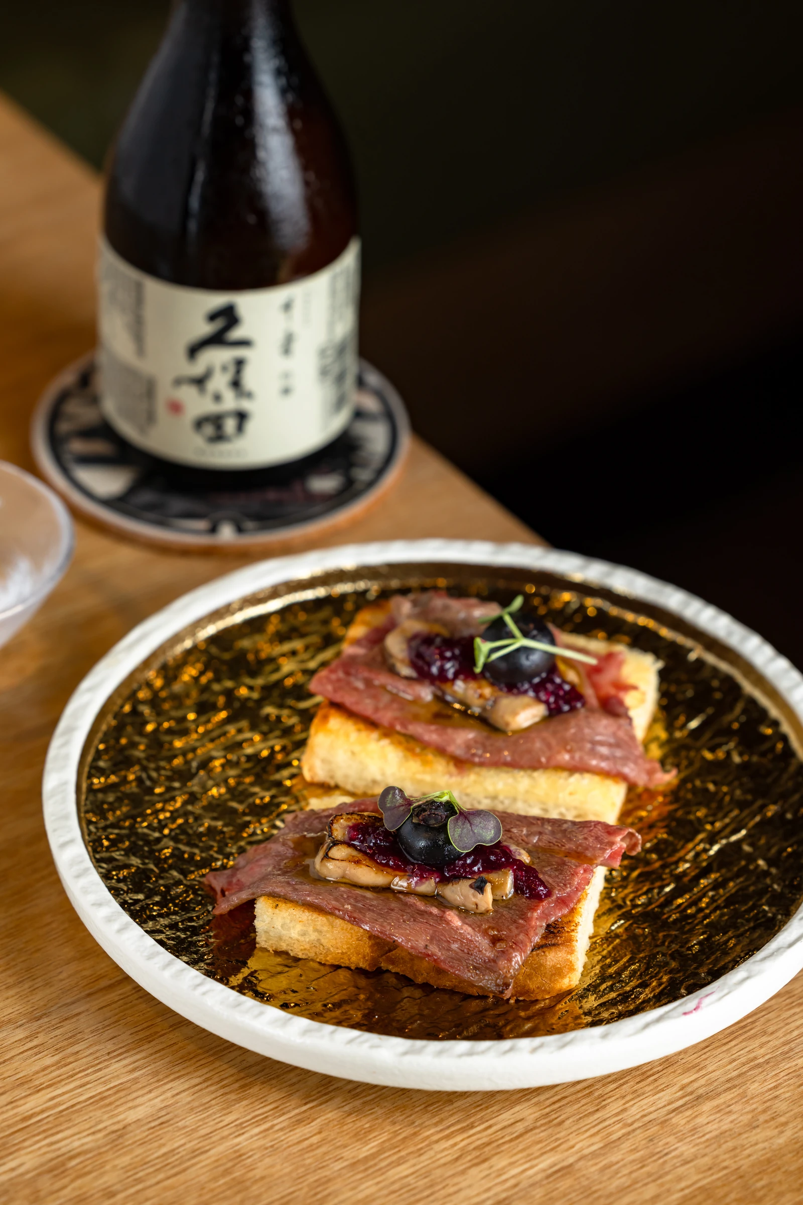 Two toast squares topped with sliced Wagyu, seared garnish and berries on a gold-toned plate